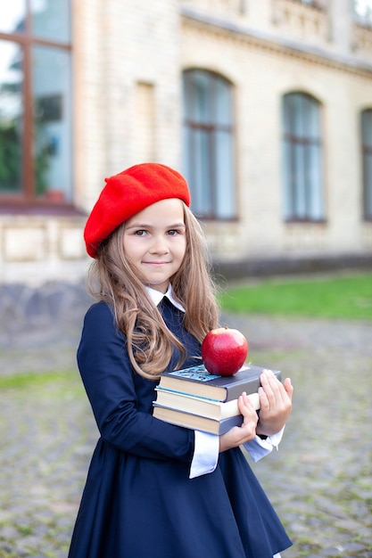 赤いベレー帽の笑顔の少女は 校舎の近くのリンゴと本の山を持っています プレミアム写真