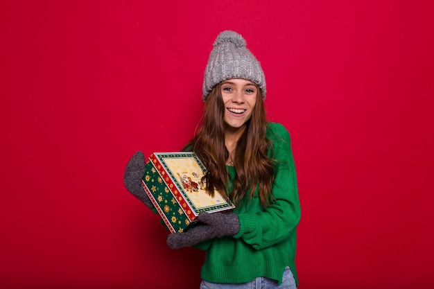 緑のセーターと赤のクリスマスプレゼントを保持している灰色のニット帽の長髪の若い女性 無料の写真
