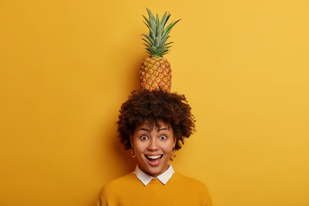 私にできることを見てください アフロの髪型を持つ陽気な面白い女性は 頭にジューシーなパイナップルを保持し 楽しさと笑いを持っています 無料の写真