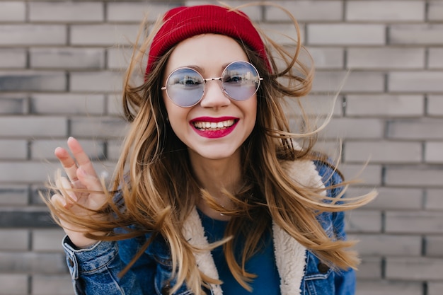 屋外の写真撮影中に浮気しているカジュアルな青い眼鏡の愛らしい女性 レンガの壁にポーズをとって明るい化粧で興奮したブロンドの女の子の肖像画 無料の写真