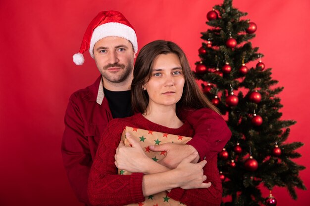 Premium Photo Lovely Cute Couple Posing At Home Infront Of Xmas Tree Celebrating New Year Man In Santa Hat And Young Woman With Christmas Gift On Red Background