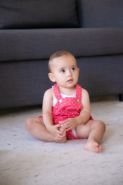リビングルームで裸足でカーペットの上に座っている素敵な小さな赤ちゃん 赤いダンガリーショーツで誰かを見て 足に触れるかわいい思いやりのある女の子 週末 子供時代 在宅のコンセプト 無料の写真