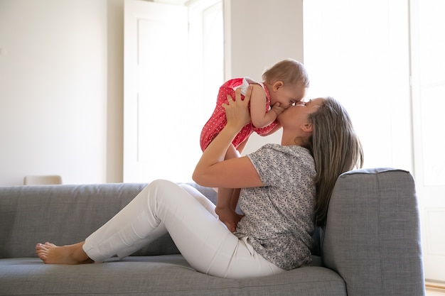 ソファに座って愛情を込めて幼い娘にキスをする愛情深い母親 手のひらで顔を閉じる幸せな女の赤ちゃん 両手で乳児を抱く長髪のお母さん 家族と母性の概念 無料の写真