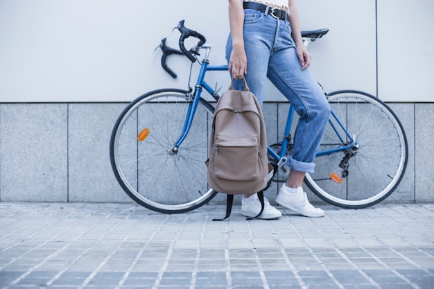 Low section of young woman standing near the bicycle holding backpack Free Photo