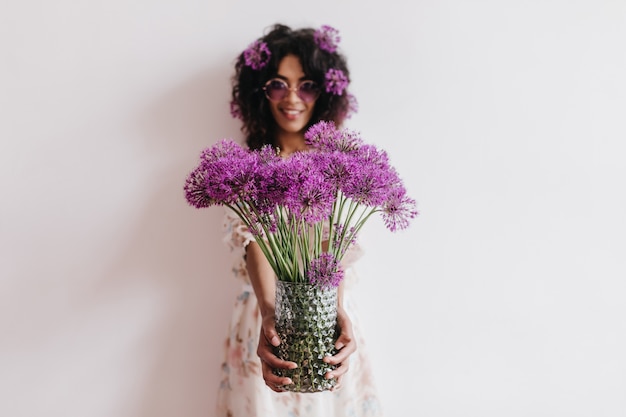 紫色の花の花瓶を持っている壮大なアフリカの女性 週末に家で楽しんでいるロマンチックな黒人若い女性の屋内の肖像画 無料の写真