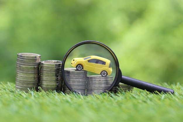 Magnifying glass with miniature yellow car model on growing stack of coins money on nature green space Premium Photo