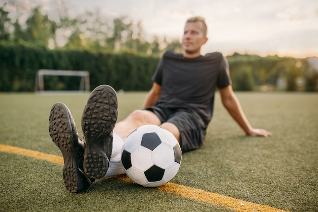 フィールドの芝生の上に座っているボールを持つ男性のサッカー選手 屋外スタジアムでのサッカー選手 試合前のトレーニング サッカートレーニング プレミアム写真