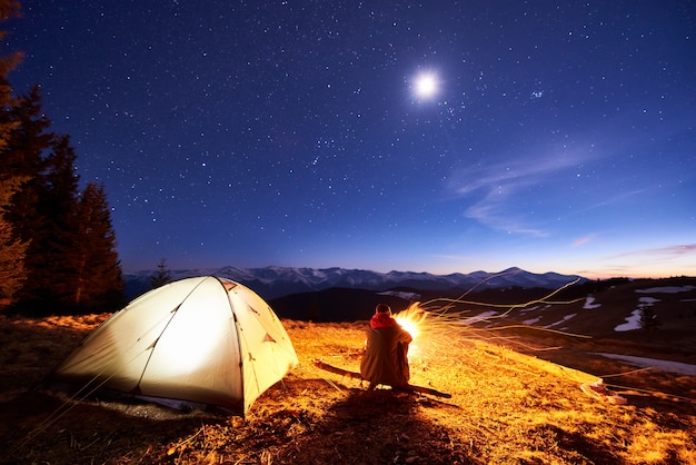 男性の観光客は夜にキャンプファイヤーの近くのキャンプで休憩し 星と月でいっぱいの美しい夜空の下でテントを張り 山の夜景を楽しんでいます プレミアム写真