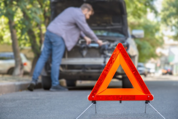 道路で車を修理する男 運転中の車の故障 ドライバーは車のボンネットの下で故障を見ています 道路上の赤い三角形の一時停止の標識 プレミアム写真