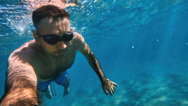 Uomo in occhiali che nuotano sotto l'acqua blu e trasparente del mar mediterraneo. tenendo la fotocamera Foto Gratuite