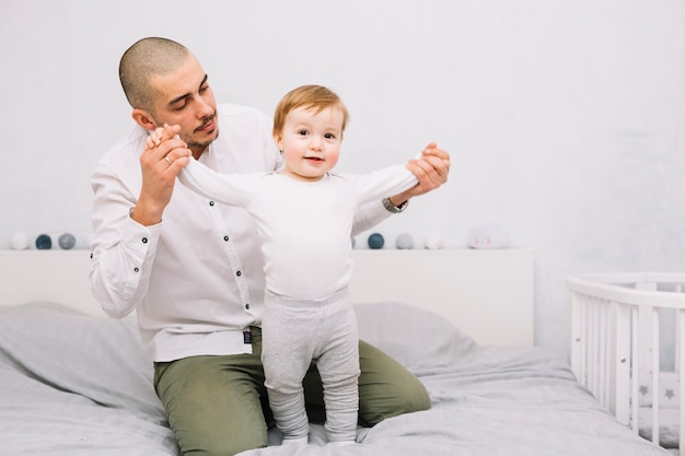 男はベッドの上に立って面白い赤ちゃんの手を繋いでいます 無料の写真