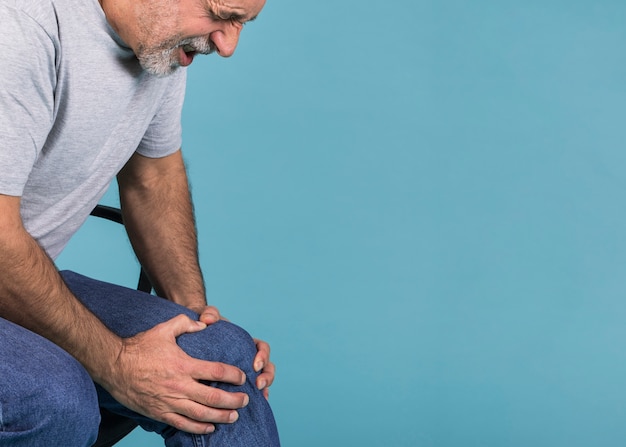 Man holding his knee in pain while sitting on chair against blue background Free Photo