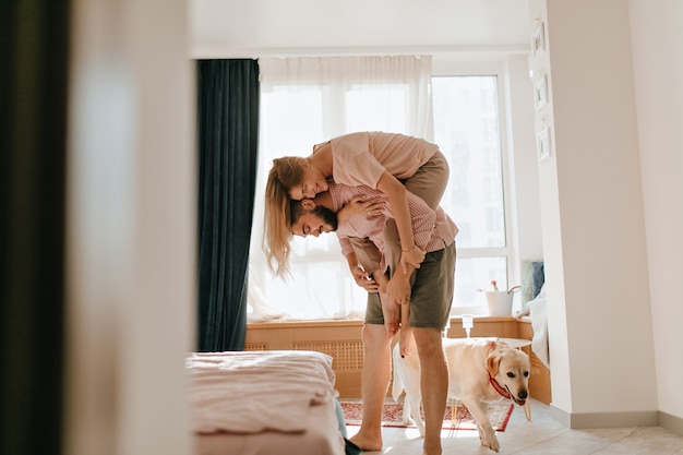 カーキ色のショートパンツを着た男性が 犬が横を歩いている間 若い妻を背負っています 恋人たちはアパートでのんびりとした週末を楽しんでいます 無料の写真