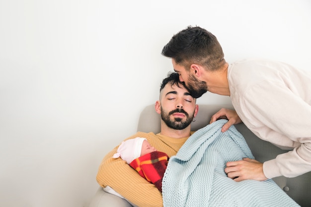 白い壁に赤ちゃんと一緒にソファで寝ている彼の彼氏の額にキスをする男 無料の写真