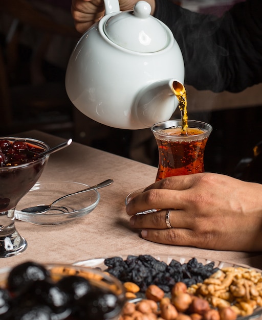 Man pouring black tea from teapot, served with dried fruits, jam Photo