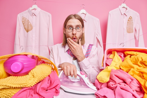 男は ピンク色の洗濯物の山に囲まれた ショックを受けたアイロンの服を見つめる 無料の写真