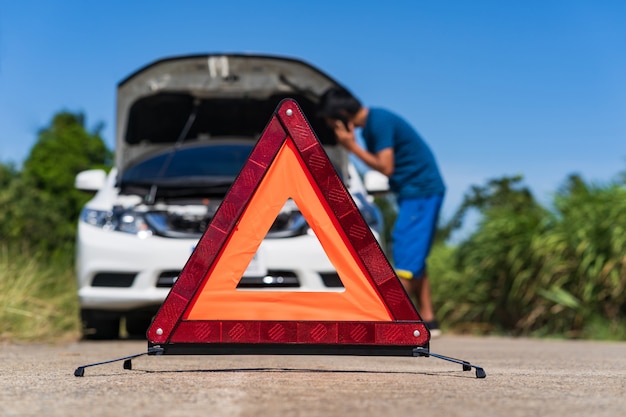 A man using a phone while having a problem car and a red triangle warning sign on the road Premium Photo