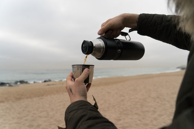 Man in warm jacket with hand tattoos, pours hot coffee or tea into thermos insulated cup on cold rainy day. Free Photo