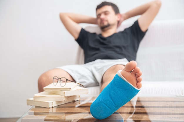 自宅のリハビリテーションで本を読んで足首の捻挫による怪我の治療のための青い副子で足を骨折した男性 無料の写真