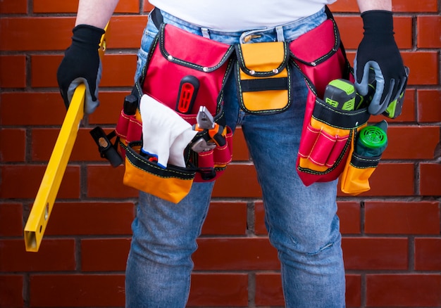 A man with a standing level in his hand against the background of a red brick wall with a full tool bag. Premium Photo
