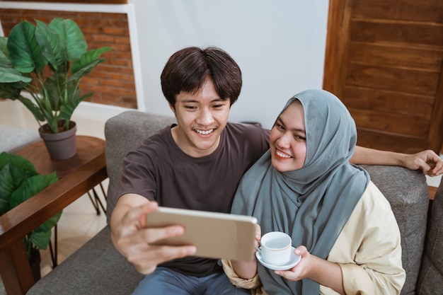 ヒジャーブの男性と女の子は 笑顔でリビングルームに座ってカップを持って自分撮りをしながらスマートフォンの画面を見ます プレミアム写真