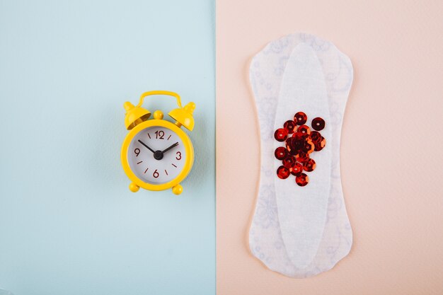 Menstruation woman hygiene concept. minimal flat lay menstrual pad and yellow alarm clock on a pink blue background. Premium Photo