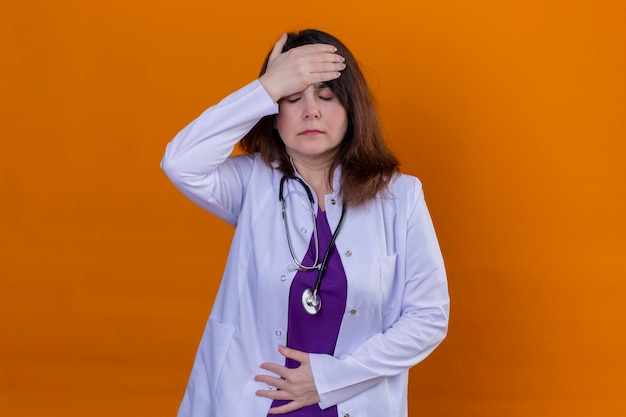 中年の女性医師が白いコートを着て、孤立したオレンジ色の背景の上に立って頭痛を持っている不健康な疲れと過労の頭に触れる聴診器で 無料の写真