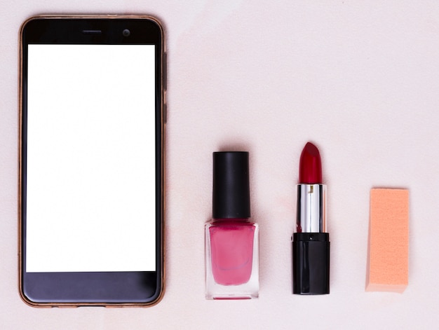 Mobile phone with white screen; nail varnish bottle; red lipstick and adhesive notes on colored backdrop Free Photo