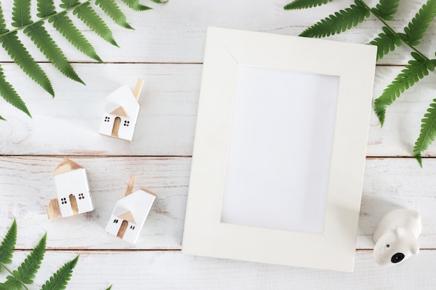 Mock up, blank white picture frame with fern leaf and miniature white house model on white wooden plank background, minimalist Premium Photo