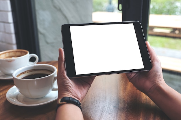 Mockup image of hands holding black tablet pc with white blank screen and coffee cups Premium Photo