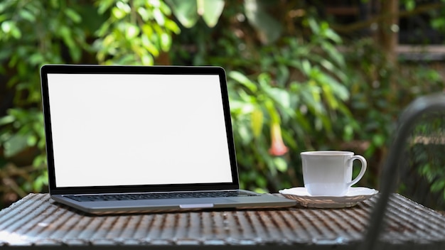  Mockup laptop blank screen and white coffee mug on iron table in the garden,green tree background.