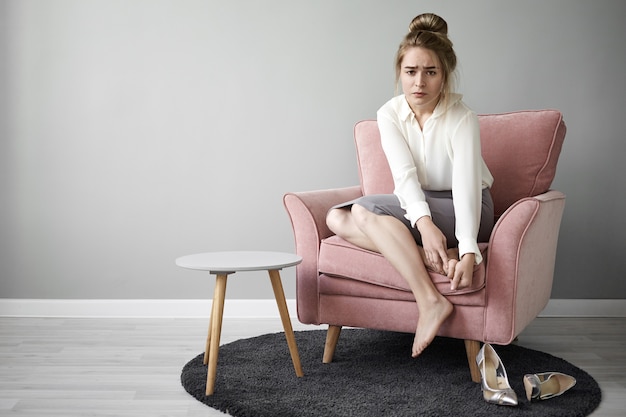 Modern hardworking young female feeling tired after long walk on high heeled shoes, sitting barefooted in armchair, massaging her feet, having painful frustrated facial expression. health and wellness Free Photo