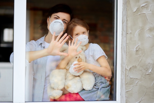 Mother with daughter child in mask looking from window, coronavirus. Premium Photo