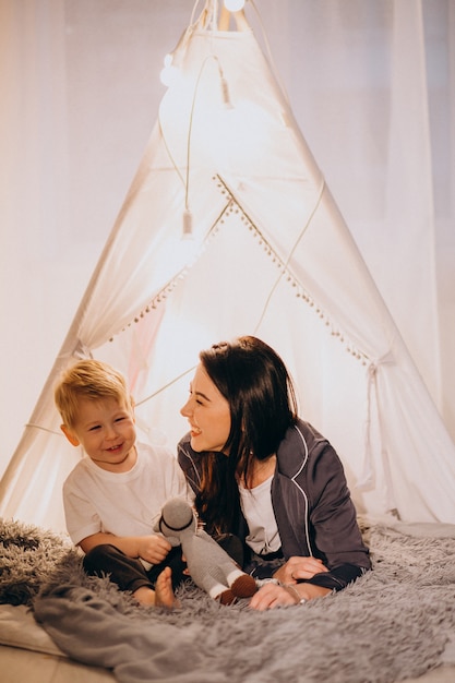 Mother With Son Sitting In Cozy Tent With Lights At Home On