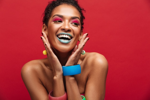 Multicolor cheerful african american woman with fashion makeup smiling and looking aside, isolated over red wall Free Photo