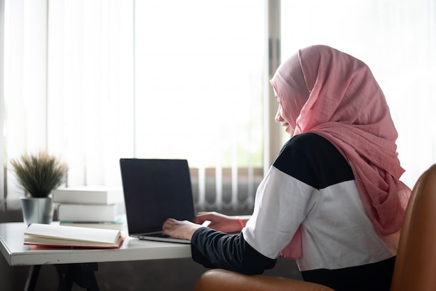 The muslim woman sitting on wooden chair,, doing work at home, using laptop and books on wooden desk, beside window, Premium Photo