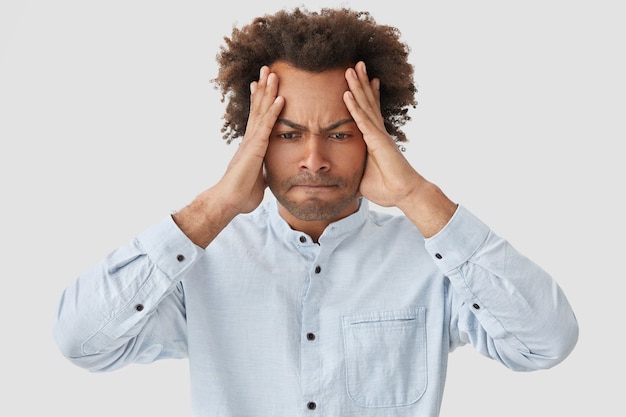Negative facial expressions concept. stressed african american male keeps hands on head, presses lips and looks desperately down, dressed in elegant-shirt, being in panic, suffers from headache Free Photo