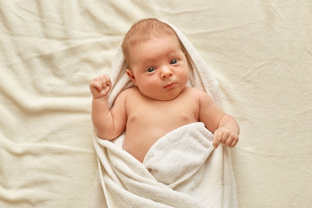 入浴後にタオルを身に着けている生まれたばかりの赤ちゃん 白い毛布の上にベッドに横たわっているかわいい小さな子供は 外のことを見て勉強しています 子供の頃と赤ちゃんの世話 無料の写真