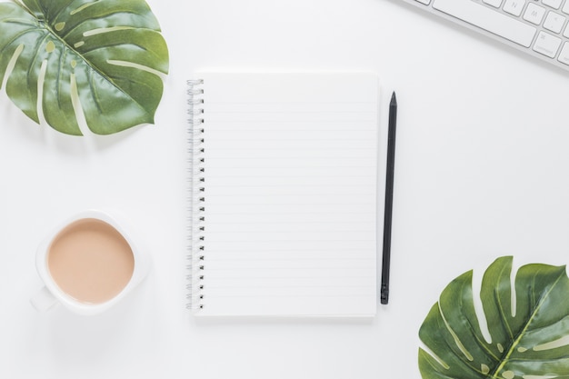 Notebook near coffee cup and keyboard on table with green leaves Free Photo