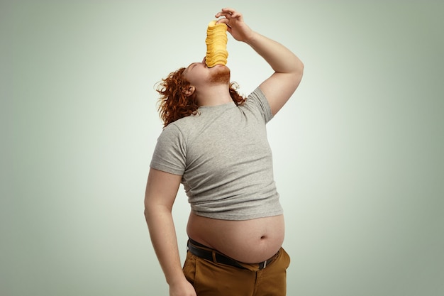 Obese overweight redhead man throwing head back, stuffing big pile of potato chips into mouth, enjoying unhealthy junk food, wearing undersized t-shirt with belly sticking out Free Photo