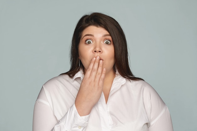 Omg. amazed emotional plus size woman in white shirt opening eyes widely and covering mouth, staring in full disbelief, holding breath, can't say anything because of shock Free Photo