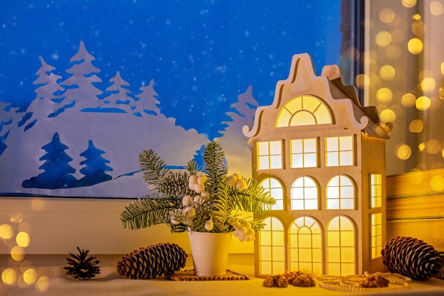 窓辺のクリスマスツリーの飾りの中で 夜の雪の窓と紙の飾りの近くに 古いヨーロッパの家の形をした常夜灯が輝いています プレミアム写真