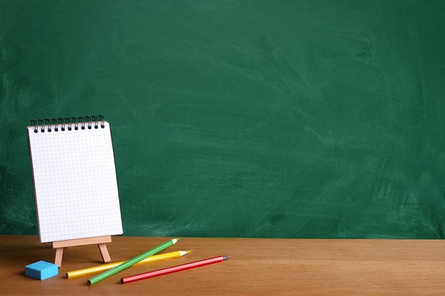 Open notebook on miniature easel and colored pencils on the background of a green chalkboard with chalk stains, copy space Premium Photo