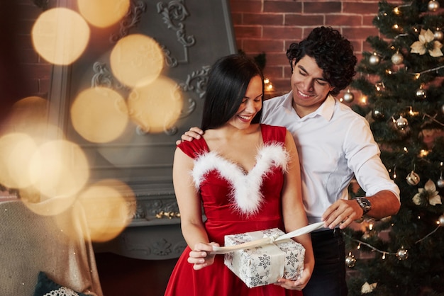 Opening the present. beautiful couple celebrating new year in the decorated room with christmas tree and fireplace behind Premium Photo
