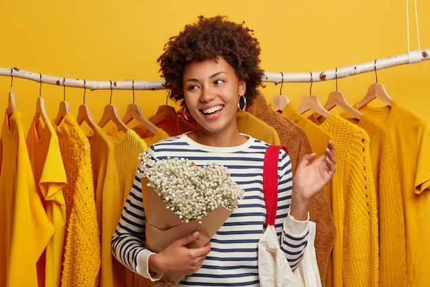 楽観的な女性のお客様は 店内での買い物に自由な時間を費やし 花束とバッグを持ってラックの黄色い服とポーズをとり 広い笑顔で脇に集中します 無料の写真