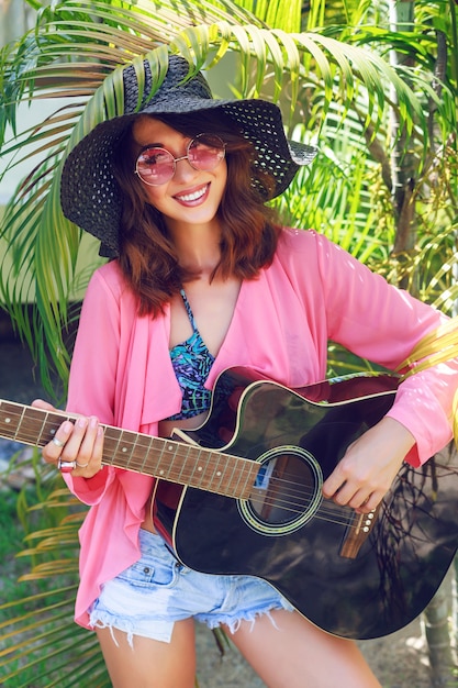 草に座って アコースティックギター を持って幸せなかなり笑顔のヒッピー女のアウトドアファッションの肖像画 熱帯の暑い国 緑の背景 帽子とピンクのサングラスの夏の服装 無料の写真