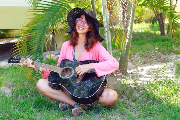 草に座って アコースティックギターを持って幸せなかなり笑顔のヒッピー女のアウトドアファッションの肖像画 熱帯の暑い国 緑の背景 帽子とピンクのサングラスの夏の服装 無料の写真
