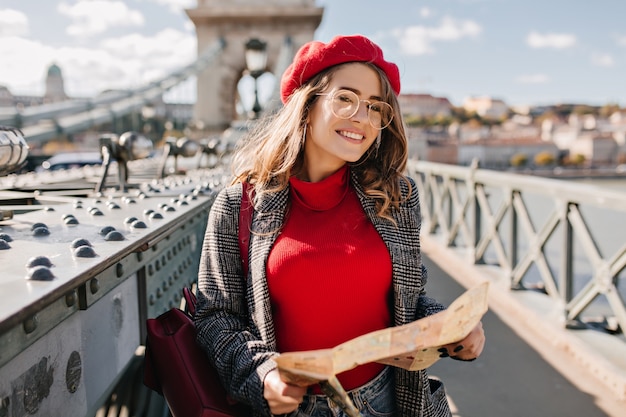 Outdoor photo of pretty female traveler in glasses holding map on the city bridge Free Photo