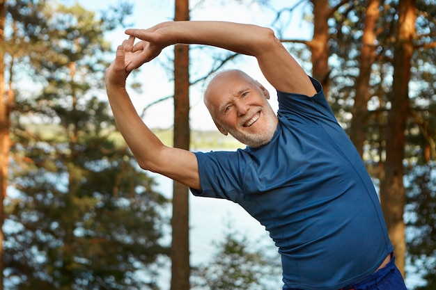 Outdoor shot of happy energetic senior retired man enjoying physical training in park, doing side bends exercise, holding hands together with broad smile, warming up body before run Free Photo