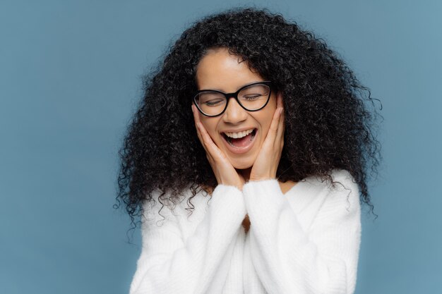 Overemotive female touches cheeks laughs from joy, keeps eyes closed, cannot stop giggling Premium Photo
